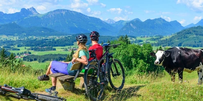 Paar mit Fahrrädern blickt auf eine malerische Berglandschaft, Kühe grasen in der Nähe im Allgäu.