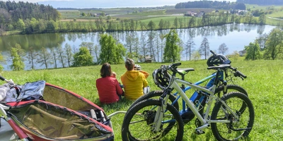 Zwei Personen sitzen am See-Ufer. Im Vordergrund befinden sich zwei Fahrränder. Eines davon hat einen Anhänger.