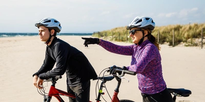 Frau am Strand zeigt auf etwas, während ein Mann nachdenklich an seinem ATB-Bike lehnt