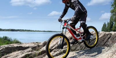 Dämpfer Fahrrad einstellen: MTB Fahrwerk optimieren Mountainbiker auf Felsen mit gelben Reifen, im Hintergrund ein Gewässer und blauer Himmel.