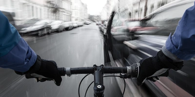 Dooring: Was ist das und wie schützt du dich vor Dooring-Unfällen beim Fahrradfahren? Blick von einem Fahrradlenker auf eine verregnete Straße mit vorbeirasenden Autos.