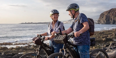 Älteres Paar mit E-Bikes an der Küste, lächelnd und im Gespräch.