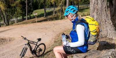 Frau sitzt. Trägt Sportkleidung und einen Fahrradhelm. Im Hintergrund steht ein E-Bike.