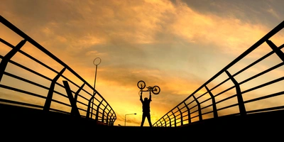 Mann auf einer Brücke hält ein Faltrad in die Luft im Sonnenuntergang