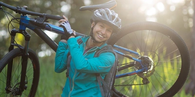 Muskeln durch Fahrrad fahren? Hier erfährst du, wie das klappt. Lächelnde Person trägt ein Mountainbike in einer sonnigen, grünen Landschaft.
