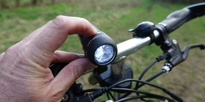 Eine Hand hält einen Frontscheinwerfer, während das Fahrrad auf einem grünen, unbefestigten Weg steht