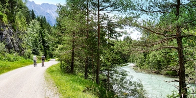 Die schönsten Flussradwege in Deutschland im Überblick Radfahrer auf einem Schotterweg neben einem klaren Fluss, umgeben von Bäumen und Bergen.