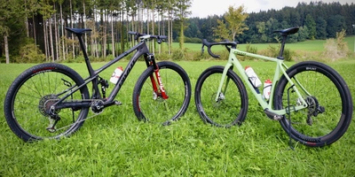 Gravelbike vs. Mountainbike Zwei Fahrräder stehen auf grünem Gras vor einem Wald, ein schwarzes und ein grünes.