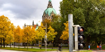 Ampel mit roten Lichtern vor herbstlichen Bäumen und historischer Architektur im Hintergrund.