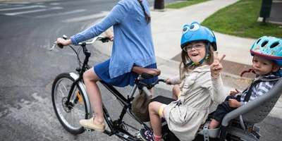 Frau fährt mit zwei Kindern auf einem Lastenrad und alle tragen einen Helm