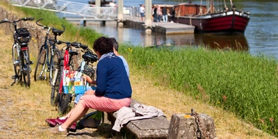 Zwei Personen sitzen auf einer Bank am Rande eines Radweges und schauen in ein Prospekt. Neben Ihnen stehen meherere Fahrräder. Am Radweg führt ein Fluss vorbei. Im Hintergrund ist ein Boot zu sehen.