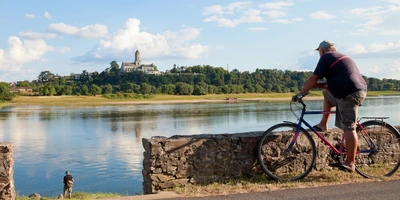 Ein Radfahrer steht an einer kniehohen Mauer und stützt seinen Fuß auf dieser ab, Blick auf den Fluss. Am Flußufer angelt ein Mann. Auf der anderen Seite des Flußes ist ein von Bäumen umgebenes Schloss und ein Dorf zu sehen.