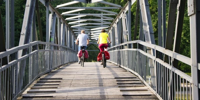 Mittellandkanal Radweg Zwei Radfahrer überqueren eine Holzbrücke in einer grünen Umgebung.