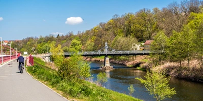 Ein Radfahrer fährt an einem Fluss entlang, mit einer Brücke und grünen Bäumen im Hintergrund.