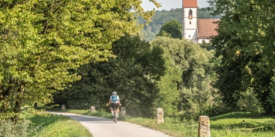 Radfahrer auf einem schmalen Weg, umgeben von Bäumen und einer Kirche im Hintergrund.
