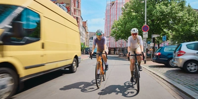 Zwei Radfahrer fahren auf einer Straße, während ein gelber Transporter vorbeifährt.
