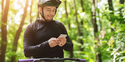 Mann mit Helm auf Fahrrad im Wald hält Handy in der Hand und schaut darauf