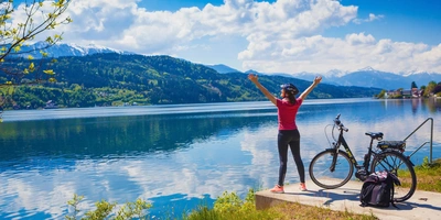 Frau steht vor einem Österreicher See neben einem Fahrrad und hebt ihre Arme freudig nach oben