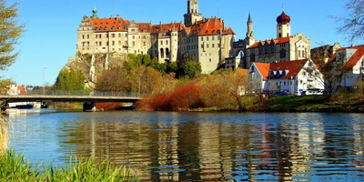 Es ist ein Fluss zu sehen. Im Vordergrund ist das grüne Ufer erkennbar. Auf der anderen Seite des Flusses ist ein Schloss, das Schloss Sigmaringen. Daneben sind kleine Häuser, ein paar Bäume. Eine Brücke führt über den Fluss in Richtung Schloss. Auf dem Fluss, die Donau, spiegelt sich das Schloss.