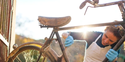 Rost am Fahrrad entfernen – so wird’s gemacht Frau reinigt ein altes Fahrrad im Sonnenlicht mit blauen Handschuhen.