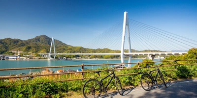 Shimanami Kaido – alles über den berühmten Radweg Zwei Fahrräder stehen auf einem Weg, mit einer Brücke und Wasser im Hintergrund.