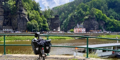 Fahrrad mit Gepäck nahe einem Fluss, umgeben von grünen Bergen und einem kleinen Ort.