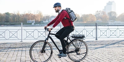 Radfahrer mit Rucksack fährt in städischer Kulisse neben einem Fluss.