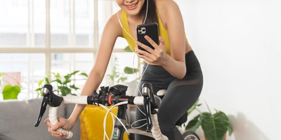 Frau in Sportkleidung lächelt beim Radfahren in ihrem Wohnzimmer und nutzt ihr Handy.