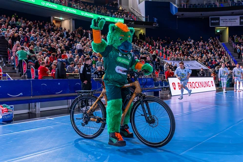 Ein grüner Bärenmaskottchen auf einem Fahrrad in einer Sporthalle, jubelnd vor jubelndem Publikum.