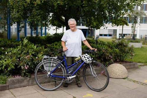 Seniorin lächelt neben ihrem blauen Fahrrad in einem begrünten Innenhof.