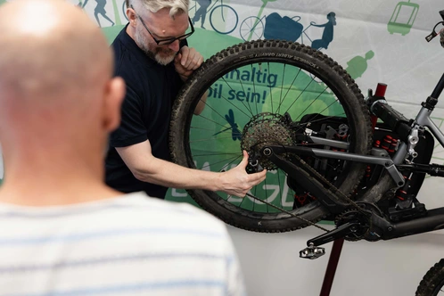 Ein Mann repariert ein Fahrrad, während ein anderer Zuschauer interessiert zuschaut.