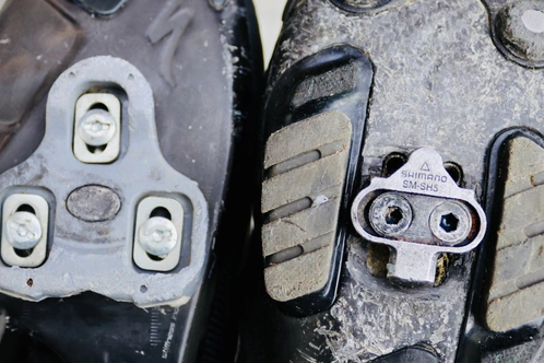 Zwei Fahrradschuhe mit unterschiedlichen Cleats und abgenutzten Sohlen.